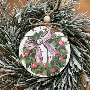 Könnte beinhalten: Ein runder, weißer Weihnachtsbaumschmuck aus Holz mit einem gemalten grünen Stechpalmenkranz, einer rosa Schleife und roten Beeren. Der Schmuck hat einen rustikalen, ländlichen Stil.