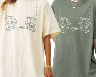 May include: Two oversized t-shirts, one ivory and one moss green, each featuring a seashell bra graphic with the text "Olivia's Bachelorette Puerto Rico July 2023" in a whimsical font. The shirts are casual and comfortable.