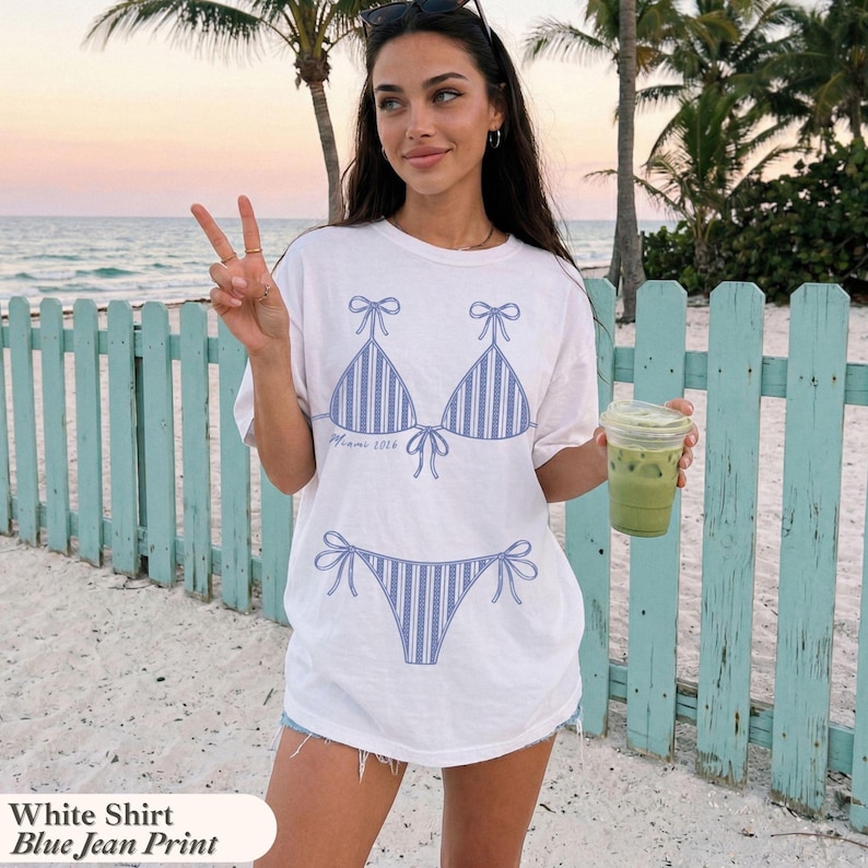 May include: A white short-sleeve t-shirt featuring a blue striped bikini graphic on the front. The bikini top and bottom are illustrated with vertical blue lines and tie-side details. The text Miami 2024 is printed in small blue script below the top.