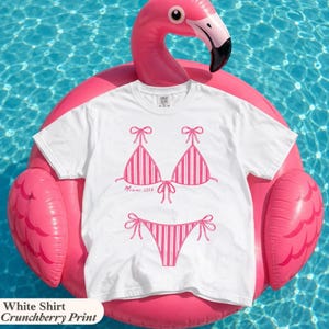 May include: A white short-sleeved t-shirt featuring a pink graphic print of a striped bikini set. The bikini top includes the text Miami 2016 printed in a cursive font. The shirt is displayed on a bright pink inflatable flamingo pool float.