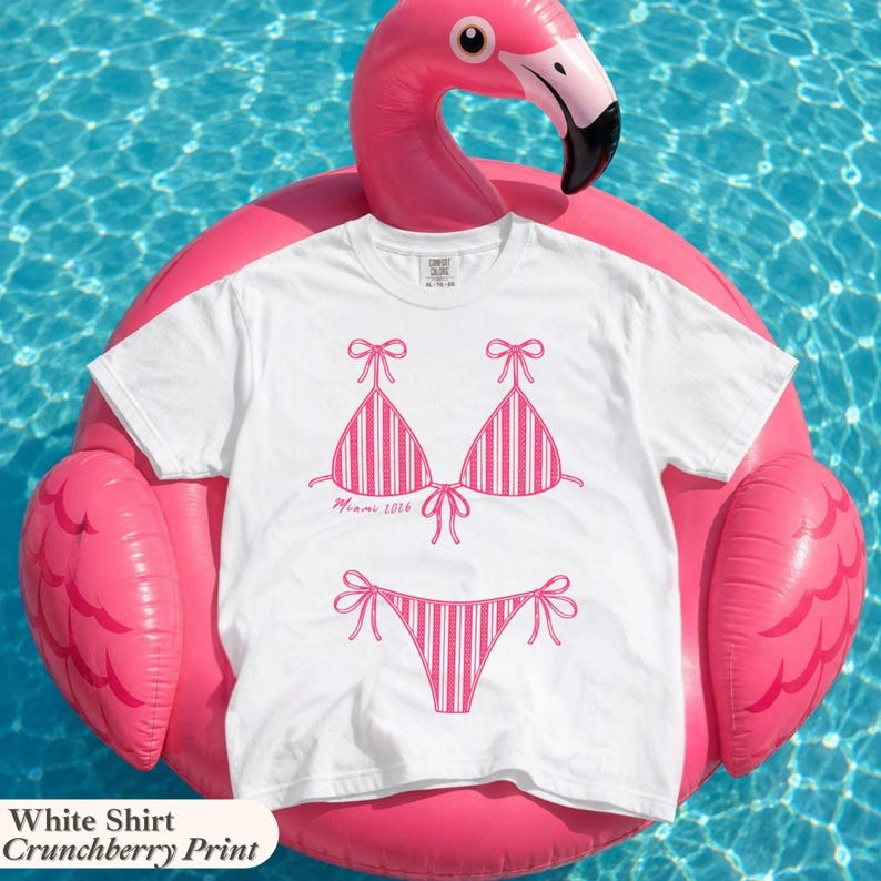 May include: A white short-sleeved t-shirt featuring a pink graphic print of a striped bikini set. The bikini top includes the text Miami 2016 printed in a cursive font. The shirt is displayed on a bright pink inflatable flamingo pool float.