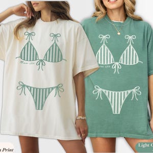 May include: An ivory t-shirt with a light green striped bikini graphic and a light green t-shirt with an ivory striped bikini graphic. Both shirts feature the text Miami 2024 printed in a small script font near the bikini top graphic.