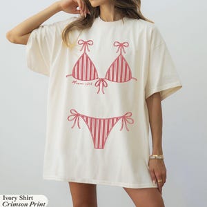 May include: An ivory short-sleeve t-shirt featuring a crimson red graphic print of a two-piece bikini with striped detailing. The text "Miami 2026" is printed in a cursive font below the bikini top. The shirt has a crew neckline and loose fit.