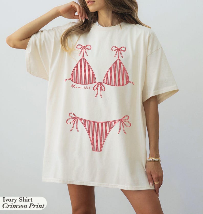 May include: An ivory short-sleeve t-shirt featuring a crimson red graphic print of a two-piece bikini with striped detailing. The text "Miami 2026" is printed in a cursive font below the bikini top. The shirt has a crew neckline and loose fit.