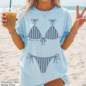 May include: A light blue t-shirt featuring a dark blue line art graphic of a striped bikini set. The graphic includes the text "Miami 2026" written in a cursive script between the bikini top and the bikini bottom pieces on the front of the shirt.
