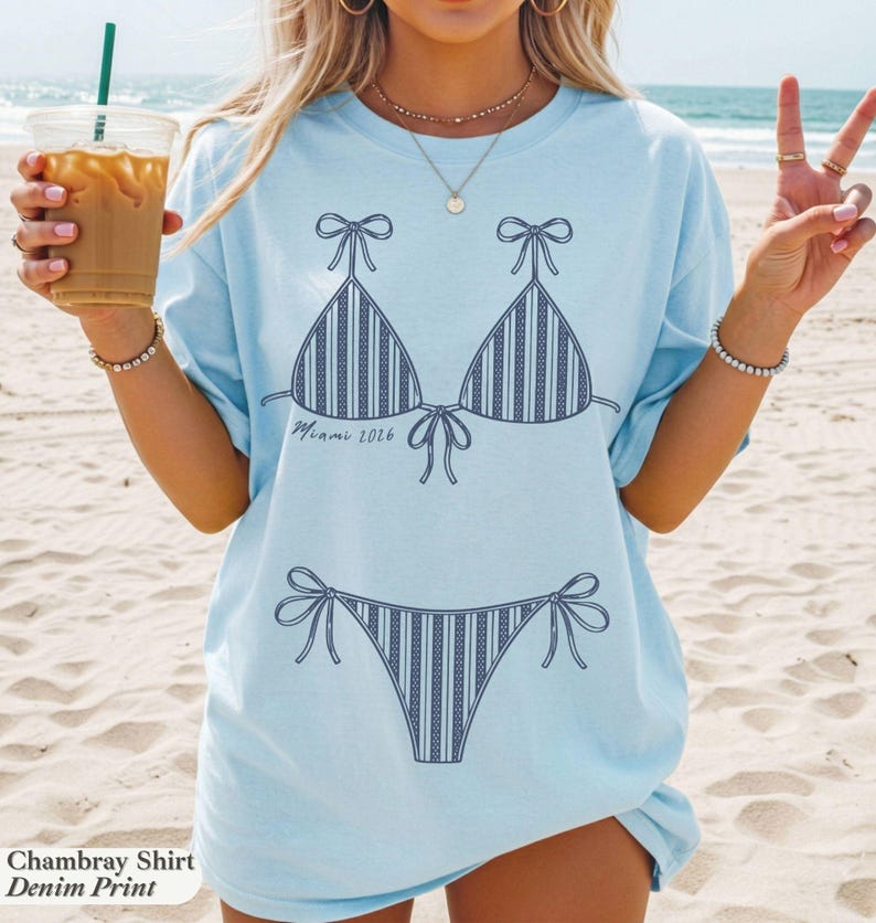 May include: A light blue t-shirt featuring a dark blue line art graphic of a striped bikini set. The graphic includes the text "Miami 2026" written in a cursive script between the bikini top and the bikini bottom pieces on the front of the shirt.