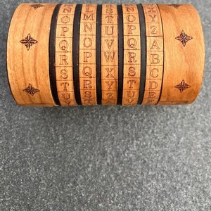 Könnte beinhalten: Ein Holzzylinder-Puzzle mit fünf drehbaren Ringen, auf denen jeweils das Alphabet aufgedruckt ist. Die Ringe sind aus hellbraunem Holz gefertigt und haben schwarze Linien, die die Buchstaben trennen. Der Zylinder ist an jedem Ende mit einem sternförmigen Design verziert.
