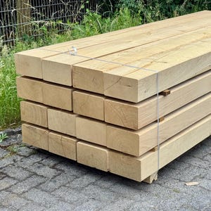 Può includere: Una pila di travi di legno di colore chiaro, probabilmente per la costruzione. Le travi rettangolari sono legate con cinghie metalliche. Il legno sembra essere legno duro. La pila poggia su un pallet.