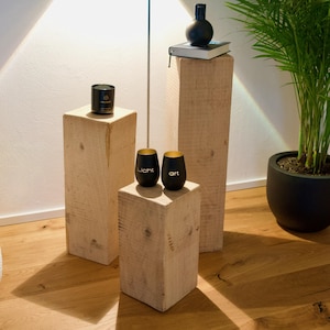 May include: Three wooden platforms of varying heights, with a black candle on the top platform, two black cups with gold rims and the words "Licht" and "Art" on the middle platform, and a black diffuser bottle on the tallest platform.