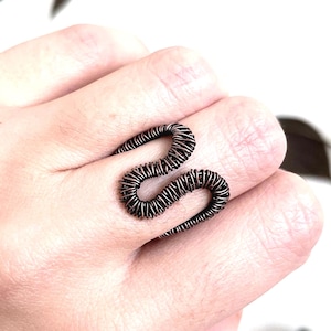 Könnte beinhalten: Ein handgefertigter, dunkelbrauner Drahtring in Form einer Schlange. Der Ring ist um einen Finger gewickelt und zeigt das komplizierte Spulendesign. Der Schlangenring ist ein einzigartiges Schmuckstück.
