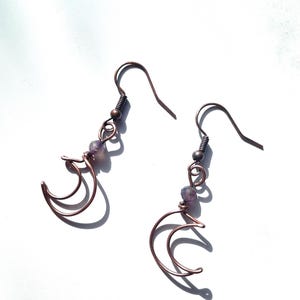 Może przedstawiać: Para kolczyków w kształcie półksiężyca w kolorze miedzi z małymi fioletowymi koralikami. Kolczyki mają druciany wzór i są prezentowane na białym tle. Haczyki są w ciemniejszym odcieniu miedzi.