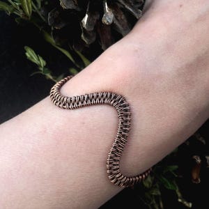 Pode incluir: Uma pulseira de arame cor de cobre com um design ondulado. A pulseira é feita de arame bem tecido, criando uma aparência texturizada. A pulseira é mostrada em um braço, com um fundo escuro.