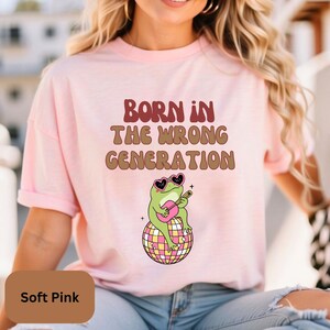 May include: A soft pink t-shirt with the text "Born in the wrong generation" in brown and red lettering. A cartoon frog wearing sunglasses is playing a guitar on a disco ball.