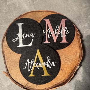 Può includere: Tre sottobicchieri rotondi in feltro nero con nomi personalizzati. Uno presenta una "L" bianca e il nome "Anna", un altro una "M" rosa e il nome "Michelle", e il terzo una "A" dorata e il nome "Alexandra".