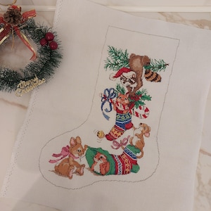 May include: A white fabric piece featuring a cross-stitch pattern of a Christmas stocking outline. The design includes a raccoon in a Santa hat, a cat, a rabbit, and a squirrel with pine branches, candy canes, and ribbons in red, green, and blue.