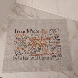 May include: A beige cross-stitch textile with the words "His Name is Jesus" in a decorative font. Other words include "Prince of Peace," "Messiah," and "Eternal Lord." The text is in shades of brown and black.