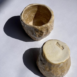Puede incluir: Dos tazas de cerámica hechas a mano con una forma orgánica y texturizada. Las tazas son de color beige cálido con sutiles motas. Una taza está boca arriba y la otra boca abajo, revelando la palabra "toula" estampada en la base.