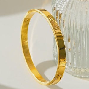 Peut inclure: Un bracelet jonc doré de forme rectangulaire. Le bracelet est doté d'une fermeture à fermoir et d'une surface lisse et polie. L'arrière-plan comprend un objet en verre transparent.
