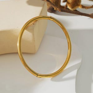 May include: A gold-coloured bangle bracelet. The bracelet is a simple, circular design with a clasp. The jewellery is displayed on a white surface with a neutral background. The bracelet is a classic piece of jewellery.