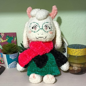 May include: A handmade plush doll of a character with white fur, pink horns, and glasses. The doll wears a red scarf, a green tunic, and a black heart-shaped emblem. The doll is approximately 10 inches tall.