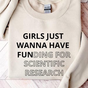 Pode incluir: Uma camisa de moletom bege com o texto "Girls Just Wanna Have Funding For Scientific Research" impresso em preto.