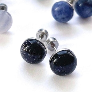 Könnte beinhalten: Eine Sammlung runder Ohrstecker mit verschiedenen Edelsteinperlen. Die Ohrringe haben Edelstahlstifte und -fassungen. Zu den Edelsteinen gehören Blau, Grün und Schwarz mit funkelnden Einschlüssen. Die Ohrringe werden auf einer weißen Oberfläche präsentiert.