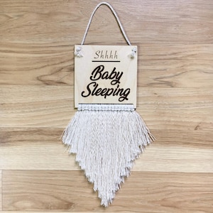 May include: A wooden sign with the text "Shhhh Baby Sleeping" in brown lettering. The sign is hanging from a white string and has a white fringe at the bottom.