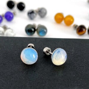 May include: A pair of round, iridescent opal-like stud earrings with silver-toned metal posts. The stones feature a smooth, domed surface with a milky, translucent appearance that shifts in color. Additional pairs of stud earrings in various colors are visible in the blurred background.