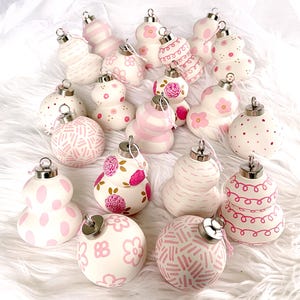 Può includere: Una collezione di 18 addobbi natalizi dipinti a mano in bianco con accenti rosa. Gli addobbi presentano vari motivi e disegni, tra cui pois, strisce, vortici e motivi floreali.
