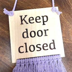 May include: A wooden sign with the text "Keep door closed" in black lettering. The sign has a lavender macrame tassel hanging from the bottom.