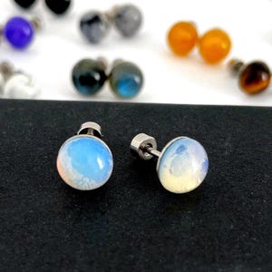 Könnte beinhalten: Zwei Opalit-Ohrstecker mit silbernen Stiften und Verschlüssen. Die Ohrringe haben einen runden, polierten Stein mit einer Mischung aus Blau-, Weiß- und Pfirsichtönen. Im Hintergrund sind weitere Ohrringe in verschiedenen Farben zu sehen.