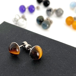 以下が含まれることがあります： 銀色の金属にセットされた、研磨された丸いタイガーアイのスタッドピアス。背景には、さまざまな色の石が付いた他のスタッドピアスがあります。イヤリングは暗い表面に表示されています。