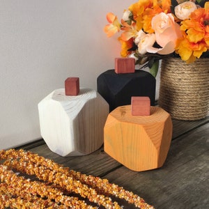 Könnte beinhalten: Drei geometrische Holz-Kürbisse in Weiß, Schwarz und Orange. Die Kürbisse sind bemalt und haben eine geometrische Form. Sie stehen auf einer Holzoberfläche mit einem Strauß orangefarbener Blumen im Hintergrund.