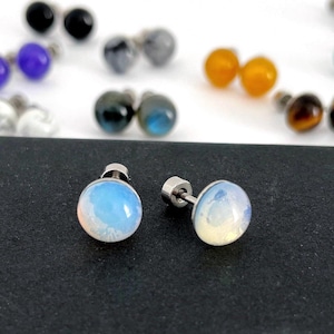 May include: A pair of round, iridescent opal-like stud earrings with silver-toned metal posts. The stones feature a smooth, domed surface with a milky, translucent appearance that shifts in color. Additional pairs of stud earrings in various colors are visible in the blurred background.