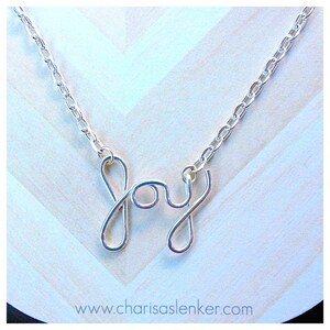 Puede incluir: Un collar de cadena de plata con un colgante de alambre de plata que dice "Alegría".