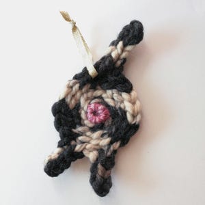 May include: A black and white crocheted ornament with a pink center, featuring a loop for hanging. The ornament is shaped like a stylized human figure.
