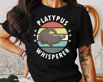May include: Black t-shirt featuring a cartoon platypus illustration inside a circular design with the text "PLATYPUS WHISPERER". The design includes a retro color scheme of red, yellow, green, and blue stripes, with white stars.