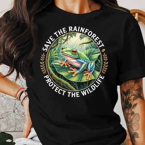 May include: Black t-shirt featuring a vibrant illustration of a tree frog on a large leaf, surrounded by lush greenery. The design includes the text "SAVE THE RAINFOREST PROTECT THE WILDLIFE" in a circular arrangement, promoting environmental awareness.