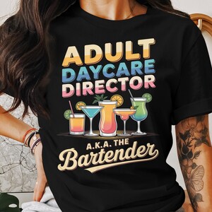 Pode incluir: Camiseta preta com o texto "ADULT DAYCARE DIRECTOR A.K.A. THE Bartender" em letras coloridas. A camiseta apresenta ilustrações de coquetéis em vários copos, com guarnições como rodelas de limão e laranja.