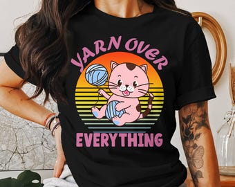 May include: Black t-shirt featuring a cartoon cat holding yarn balls. The cat is pink with brown spots, set against a sunset background. Text reads "YARN OVER EVERYTHING" in pink and white.