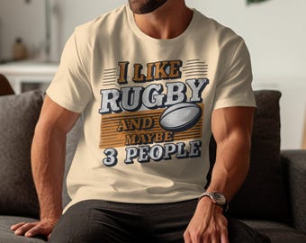 May include: A beige t-shirt with the text "I like rugby and maybe 3 people" printed on it. The text is in a brown and blue colour scheme and includes a graphic of a rugby ball.