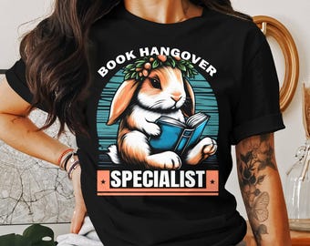May include: Black t-shirt with a graphic of a bunny wearing a flower crown and reading a book. The text "Book Hangover Specialist" is printed on the shirt.