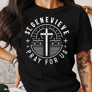 May include: Black t-shirt with a white circular design. The design includes a cross and the text "ST. GENEVIEVE PRAY FOR US". The text is in a bold font, and the design is centered. The t-shirt has a crew neck.