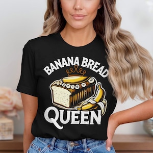 May include: Black t-shirt with a graphic of banana bread, a crown, and a banana. The text on the shirt reads "BANANA BREAD QUEEN" in white. The shirt is made of a soft material.