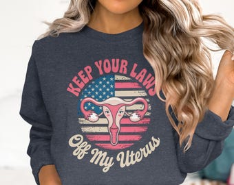 May include: Navy blue sweatshirt with a graphic design featuring a uterus illustration within a distressed American flag circle. The text reads "Keep Your Laws Off My Uterus" in pink and off-white.