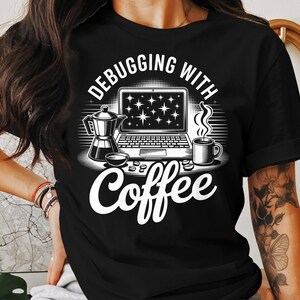 Puede incluir: Camiseta negra con un gráfico blanco. El gráfico presenta un portátil con estrellas, una cafetera, una taza de café y las palabras "Debugging with Coffee."