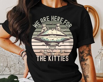 Może przedstawiać: Czarny T-shirt z retro grafiką latającego spodka z kotami. Na koszulce nadrukowany jest tekst "We are here for the kitties".