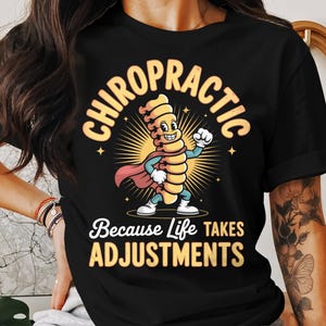 Op de afbeelding: Zwart T-shirt met een stripfiguur van een ruggengraat in een superheldenpose. De tekst luidt "CHIROPRACTIC" erboven en "Because Life Takes ADJUSTMENTS" eronder. Het ontwerp bevat een zonsopgang en sterren.