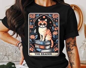 May include: Black t-shirt featuring a graphic of a cat sitting on a stack of books. The graphic is in a vintage tarot card style with the text "THE READER" at the bottom.