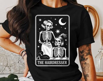 May include: Black t-shirt with a white graphic depicting two skeleton figures in a salon, with the text "THE HAIRDRESSER" beneath. The design includes a crescent moon, stars, and a stylized hair salon scene.